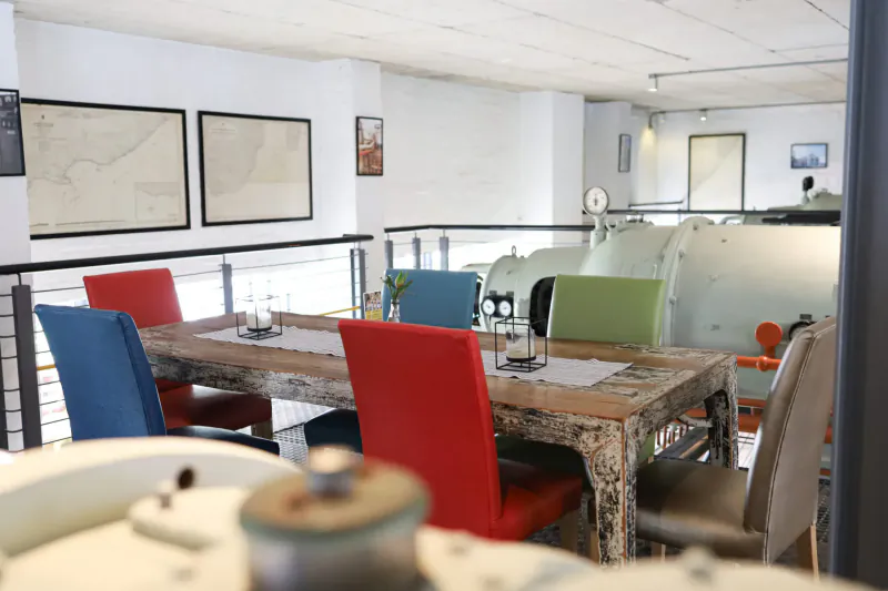Rustic wooden dining table with colorful chairs, glasses, and placemats in Turbine Hotel's modern mezzanine lounge with maps and machinery.