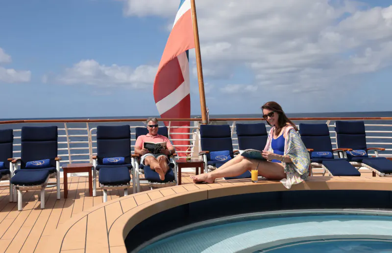 On luxury yacht deck, man in pink shirt reads on lounge chair, woman in sarong sips coffee by pool, ocean view.