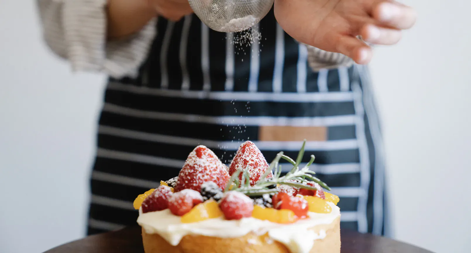 Close-up of chef in striped apron dusting powdered sugar on strawberry and mango topped cream cake