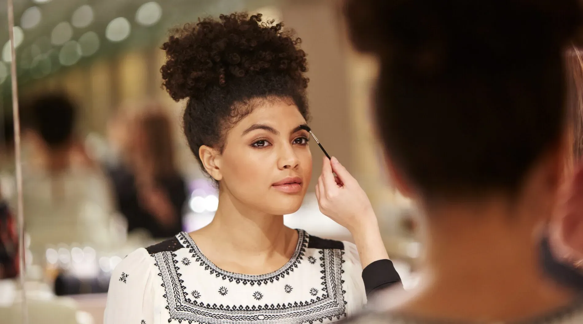 Woman with curly hair getting eyeliner applied by makeup artist in cosmetics store mirror reflection