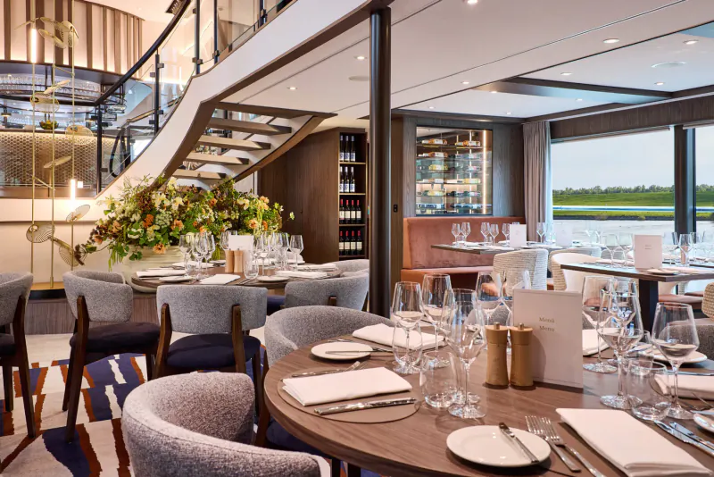 Elegant dining area on VIVA Cruises ship with curved staircase, wine racks, set tables, and river view.
