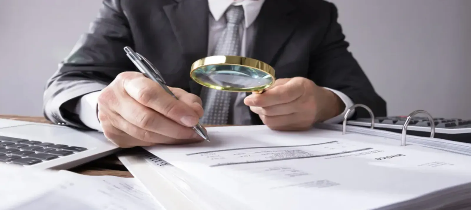Businessman in suit at desk examines documents with magnifying glass and pen, laptop nearby