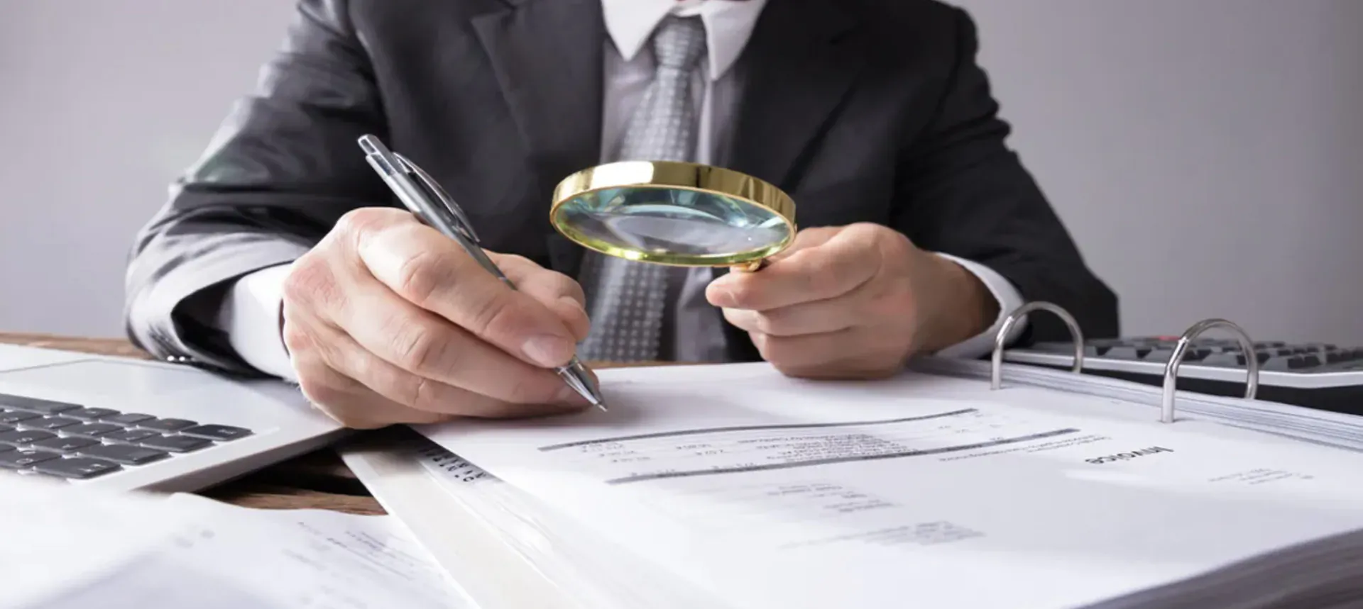 Businessman in suit at desk examines documents with magnifying glass and pen, laptop nearby