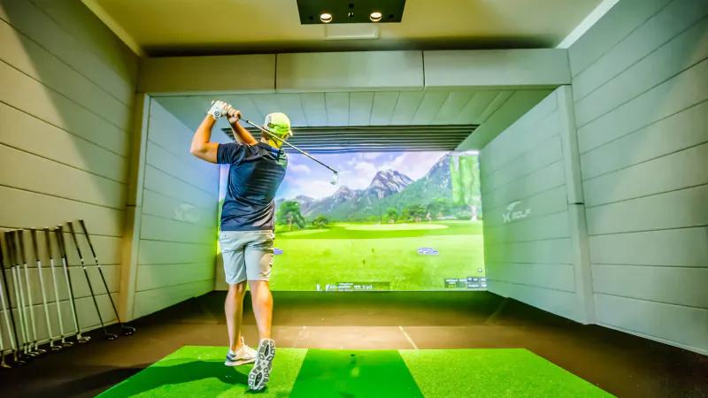Man in blue shirt and green cap swings golf club in indoor simulator with mountain golf course screen