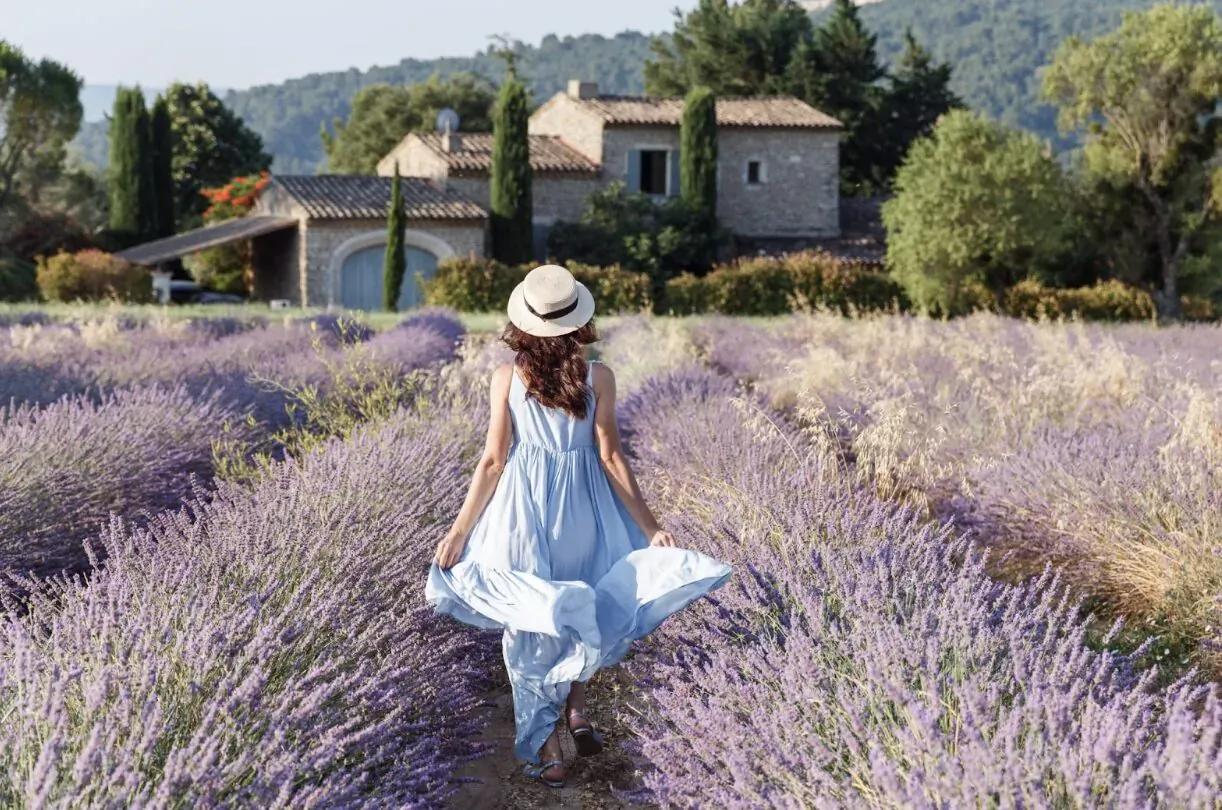 Woman in flowing blue dress and straw hat walks through Provence lavender field toward stone villa.