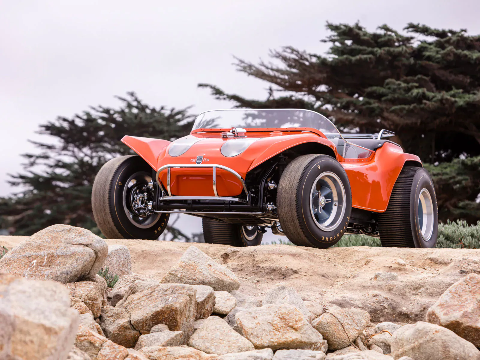 Bright orange dune buggy parked on rocky coastal bluff amid cypress trees at Concours d'Elegance
