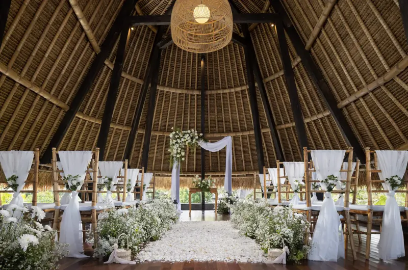 Elegant Bali wedding venue in thatched pavilion with white-draped chairs, floral arch, and petal aisle