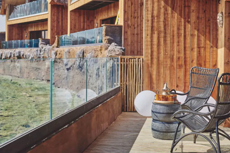 Luxury wooden safari lodge balcony with wicker chairs, barrel table, lamp, glass railing, and mountain view.
