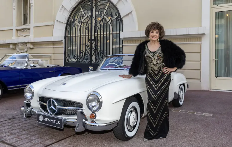 Elegant woman in black beaded dress and fur stole poses beside white Mercedes-Benz 300SL at concours d'elegance.
