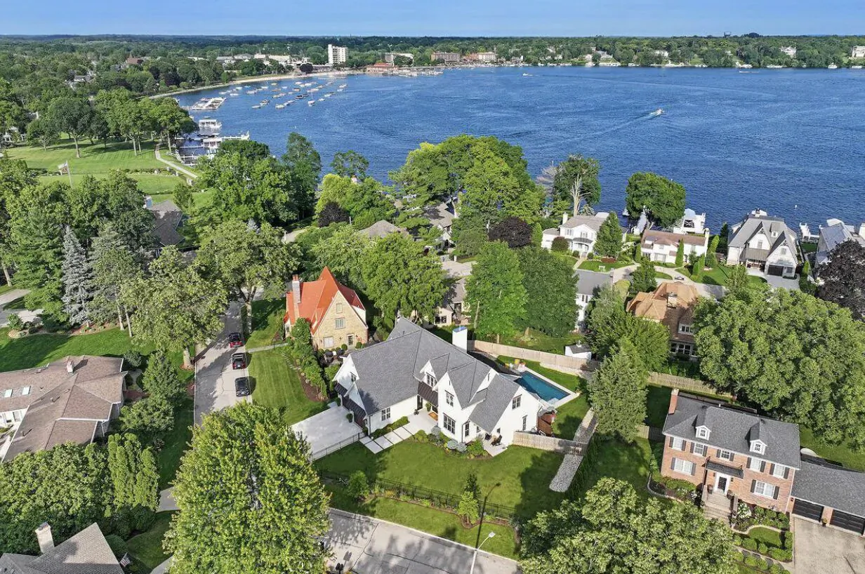 Aerial view of white waterfront mansion with pool, surrounded by green lawns, trees, neighboring homes, and lake with boats.
