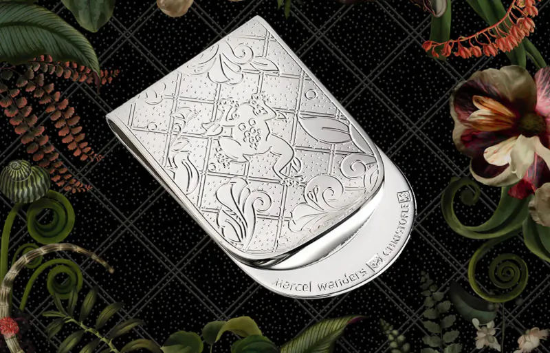 Silver money clip engraved with Jardin d’Éden motifs by Marcel Wanders for Christofle, on black floral backdrop.