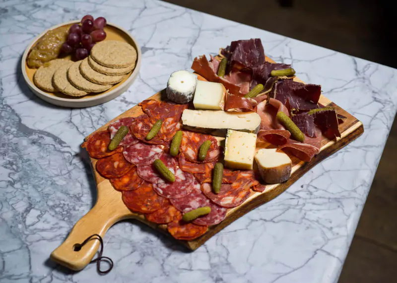 Wooden board with sliced chorizo, salami, pickles, cheeses, and crackers beside grapes and oatcakes on marble surface