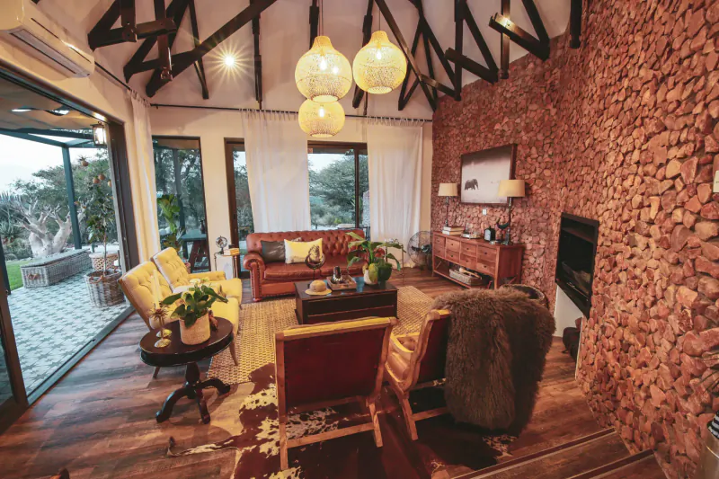 Spacious Namibian lodge living room with wooden beams, pendant lamps, leather sofas, brick walls, plants, and patio view.