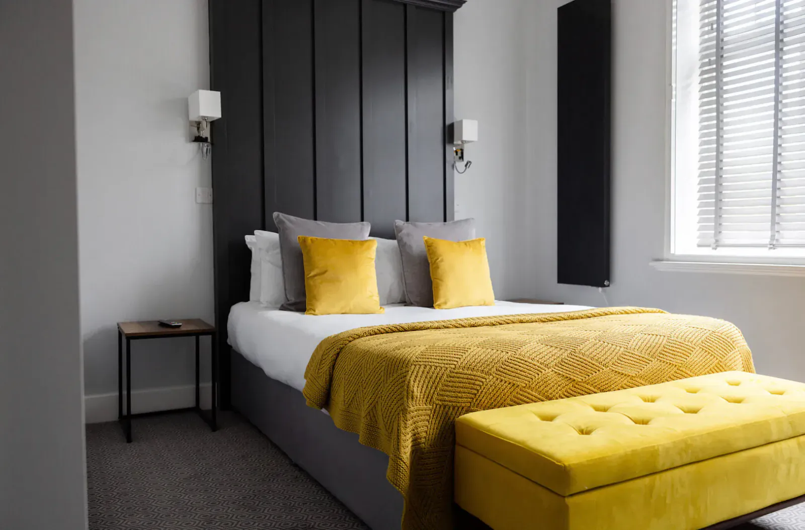 Modern hotel bedroom with black headboard, yellow pillows and throw, bedside table, and ottoman by window.