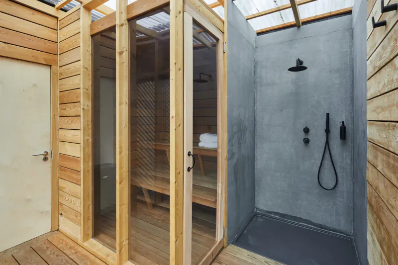 Outdoor wooden sauna with glass doors and adjacent gray-tiled shower area under a glass roof.