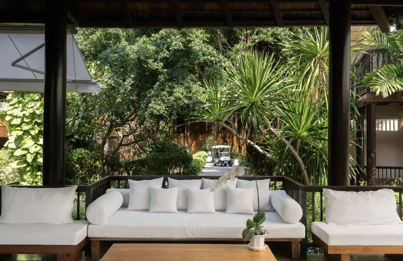 Open-air pavilion with white cushioned lounge seating, low wooden table with orchid, lush tropical garden and pool view.