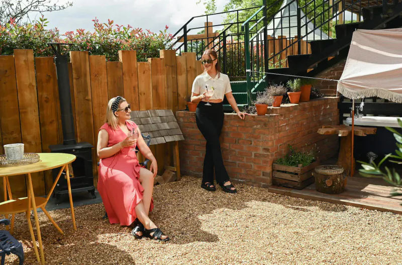 Two women chatting in stylish garden patio at The Rabbit Hotel & Retreat, one seated in pink dress holding drink, surrounded by plants and green stairs.