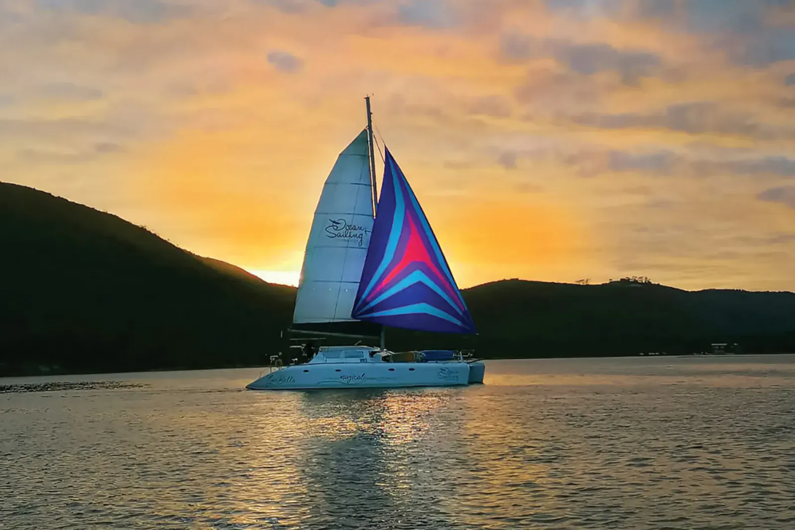 White catamaran with vibrant blue sails sailing on calm water at Knysna sunset, green hills backdrop.