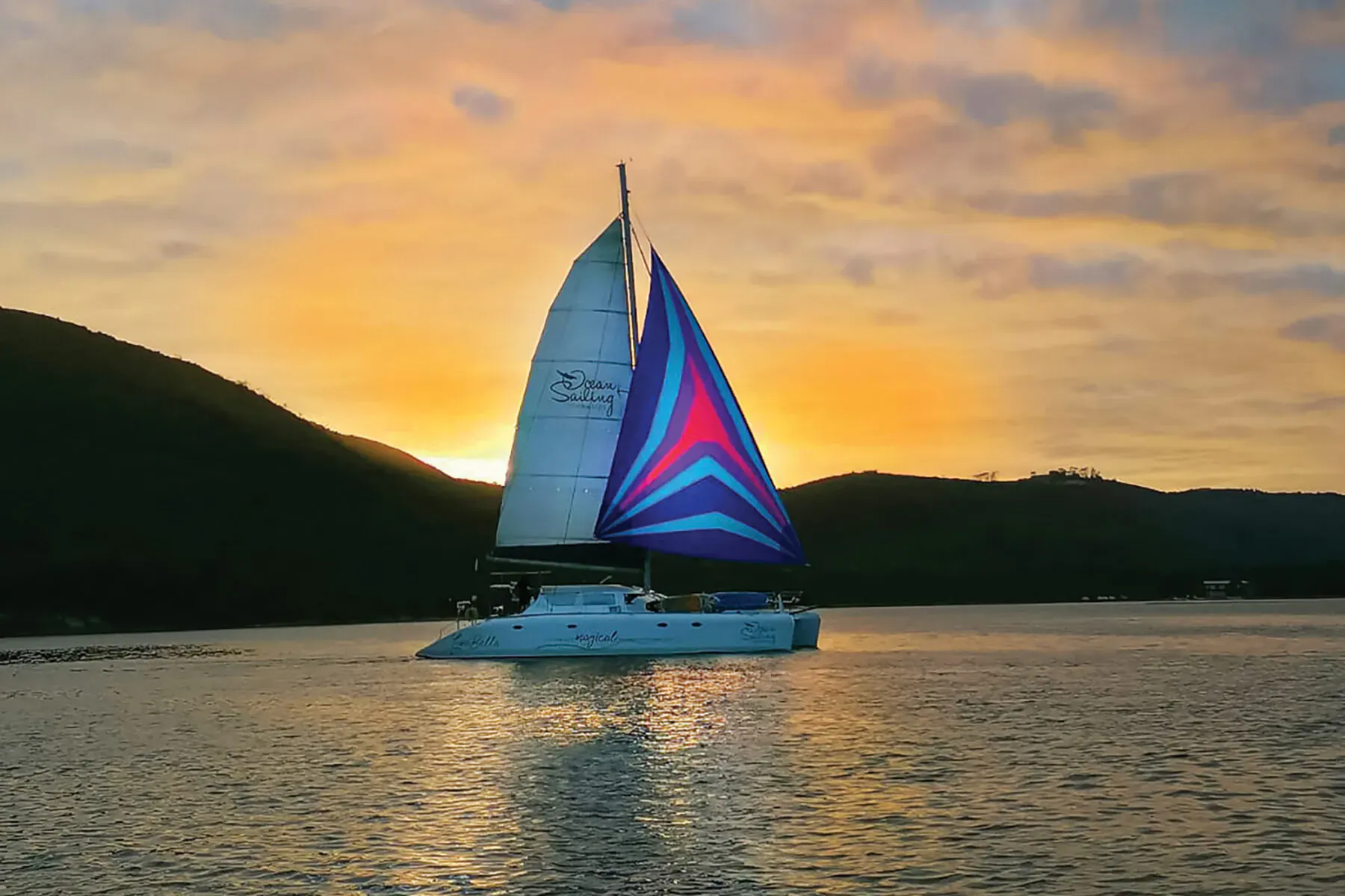 White catamaran with vibrant blue sails sailing on calm water at Knysna sunset, green hills backdrop.