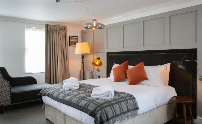 Cozy hotel room at The Black Bull with king bed, white towels, orange pillows, gray plaid throw, armchair, and pendant lamp.
