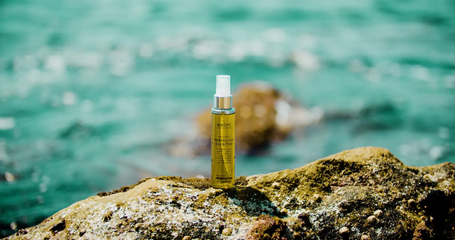 Golden oil spray bottle on rocky shore against turquoise sea, Elm Organics Norway sustainable luxury amenity