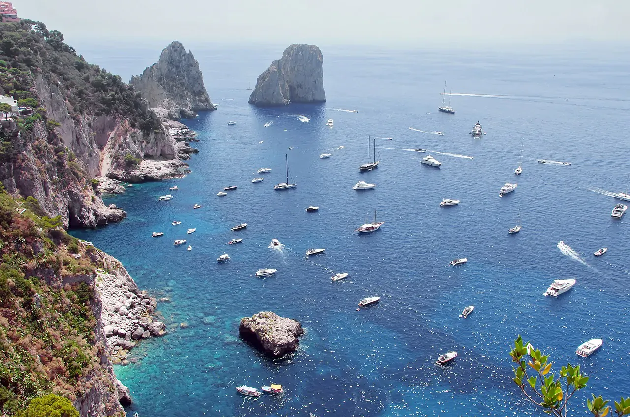 Aerial view of Capri's rugged cliffs and Faraglioni rocks rising from the turquoise sea dotted with yachts in Gulf of Naples.