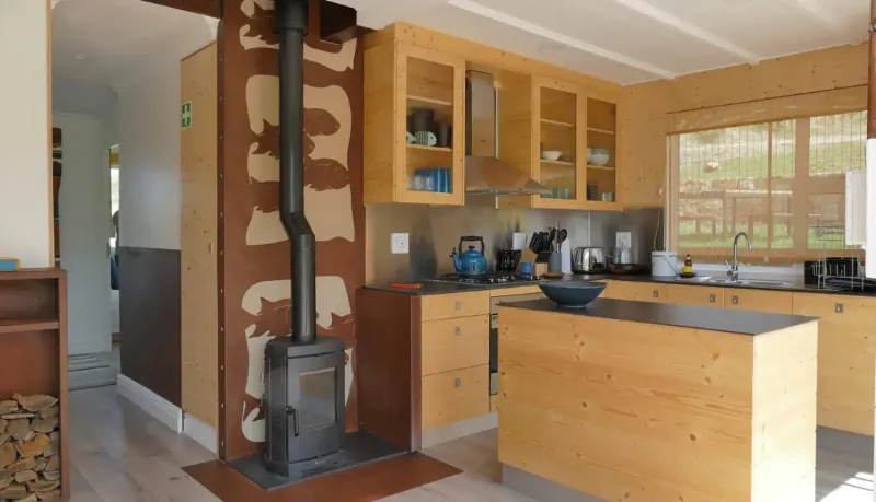 Modern wooden kitchen with black woodstove, cabinets, island counter, and valley view through windows in Elgin Valley.
