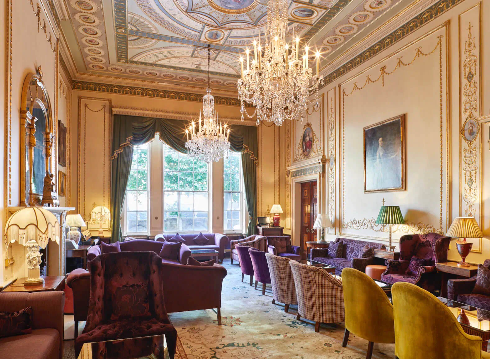 Opulent Home House lounge in London with chandelier, ornate ceiling, large windows, velvet armchairs, and lamps.