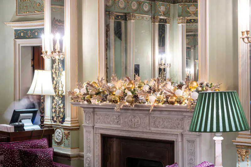 Festive gold floral garland on ornate fireplace mantel in elegant Home House room, with lamps and purple chairs.