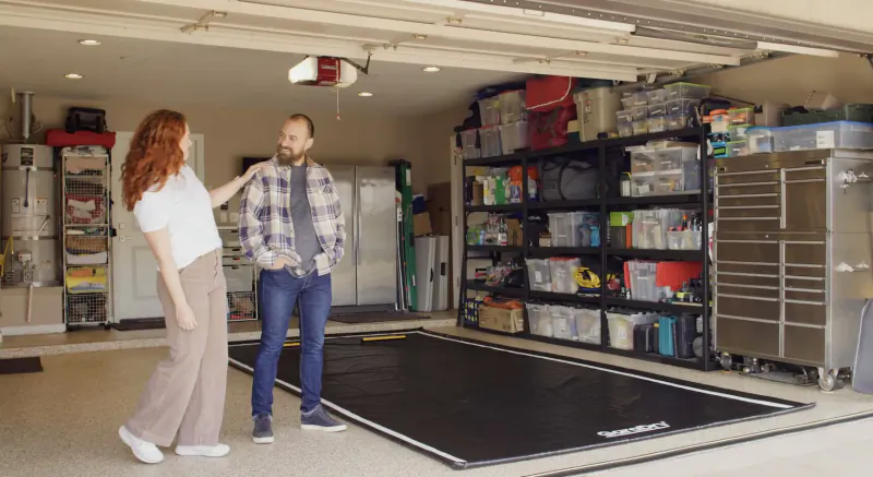 Red-haired woman and bearded man stand by GaraMat black garage floor mat amid shelves and storage.