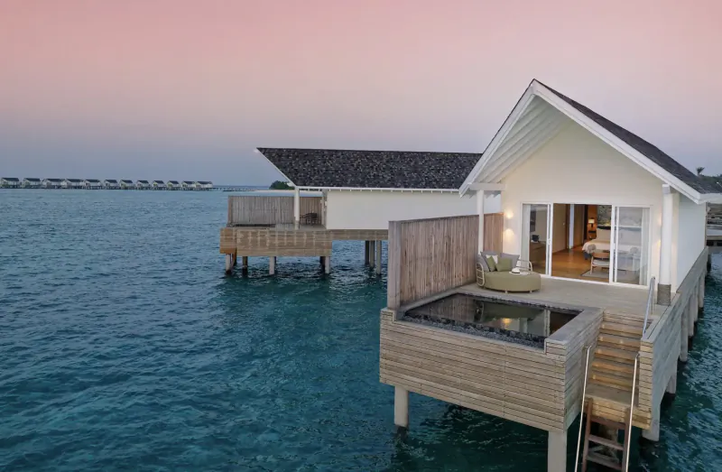 Luxurious white overwater villa with lit glass doors, jacuzzi, and steps on turquoise lagoon at sunset, Amari Raaya Maldives.