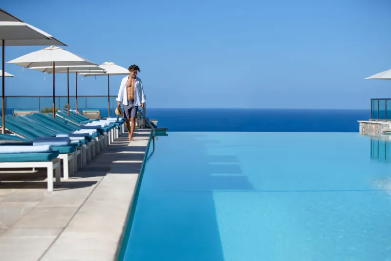 Man in white shirt and swim trunks walks by infinity pool with loungers and ocean view at Jumeirah Port Soller Hotel & Spa