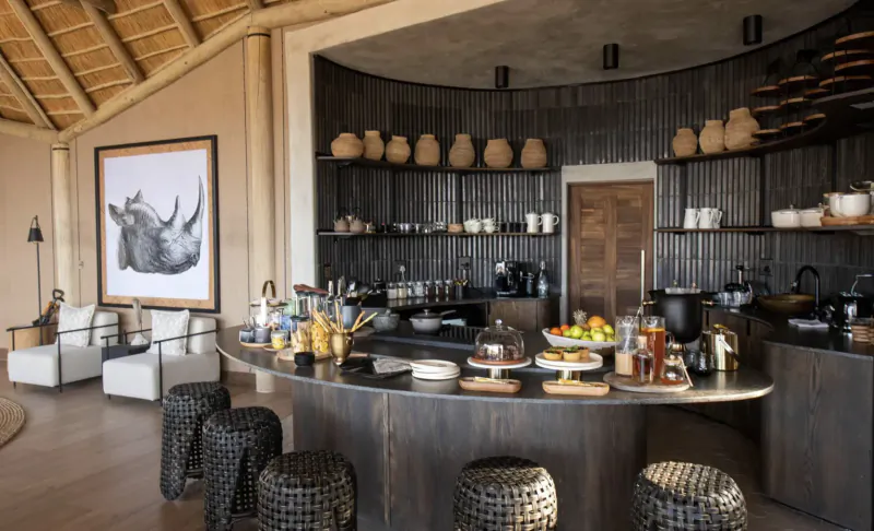 Luxurious Camp Kala kitchen bar with wooden shelves, terracotta pots, fruit, and large black rhino artwork on curved wall.