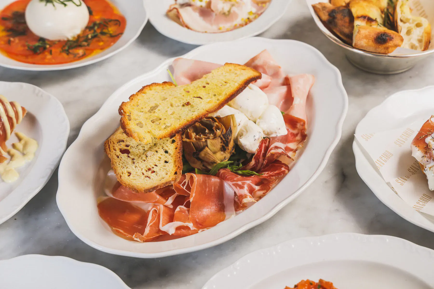 Oval plate of prosciutto, burrata, mushrooms topped with garlic bread, surrounded by Italian antipasti on marble table