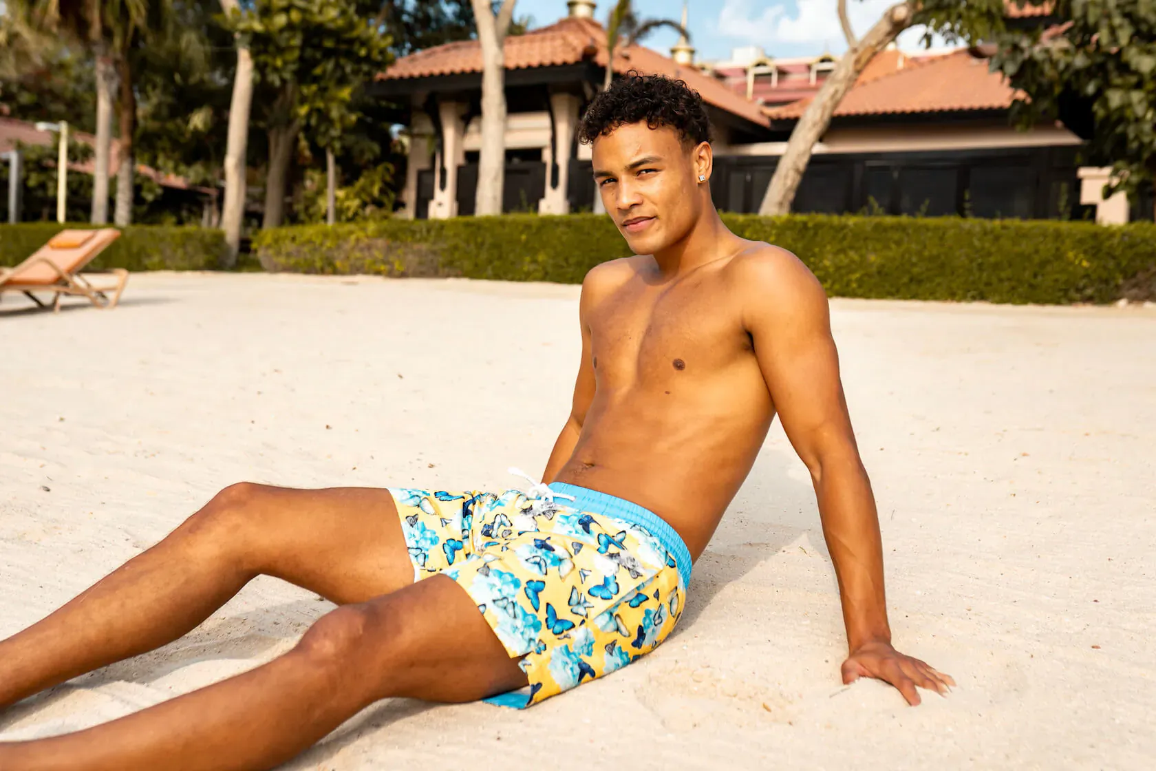 Fit mixed-race man in colorful floral swim shorts sits shirtless on sandy beach with palm trees and luxury resort behind.