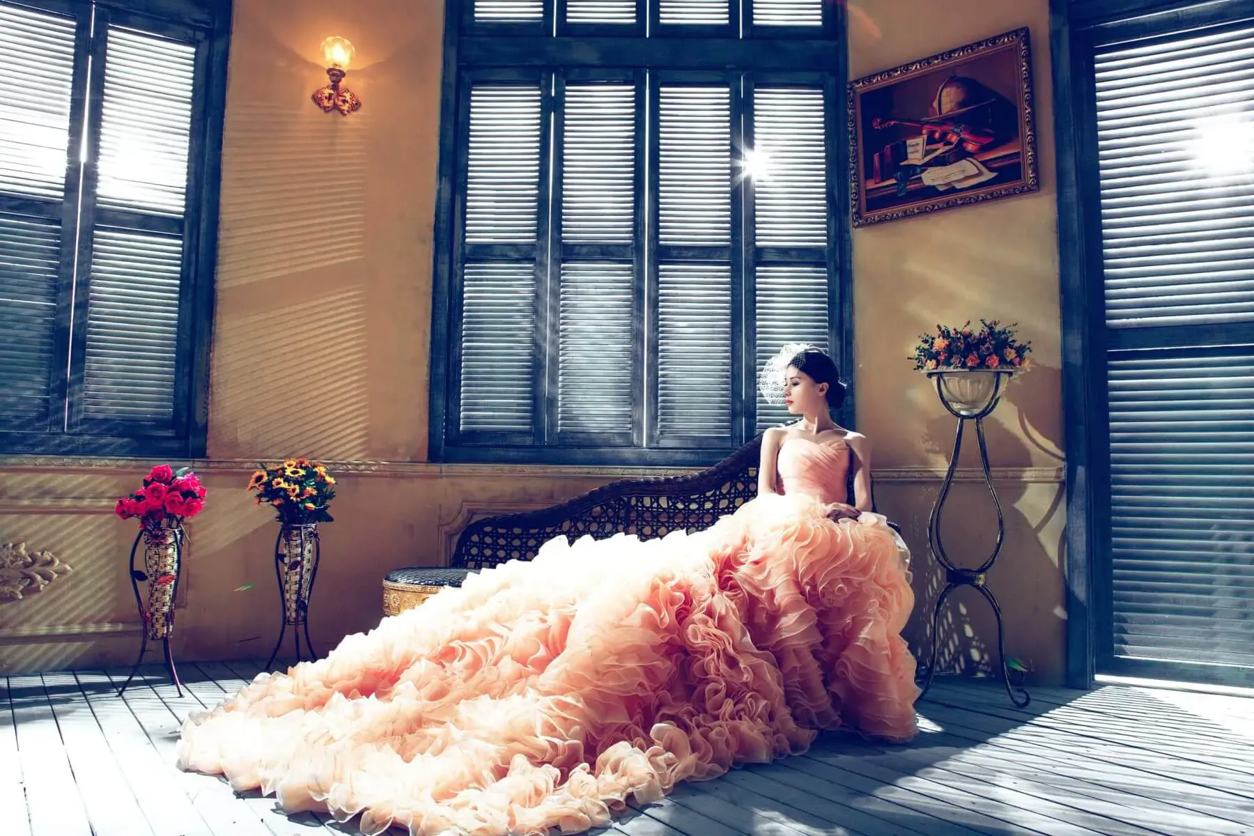 Elegant woman in flowing pink ruffled gown seated on chaise in sunlit Regency-style room with shutters and flowers