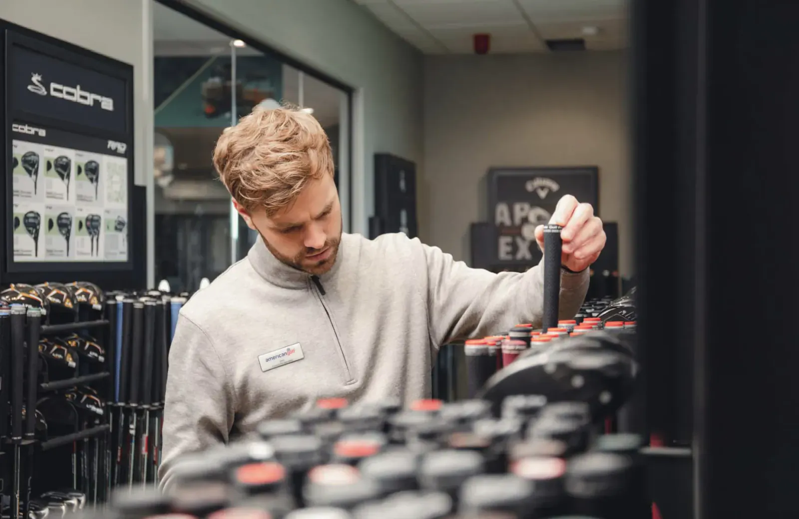 Male staff member in grey pullover selects black Cobra golf club from rack in store with TaylorMade displays