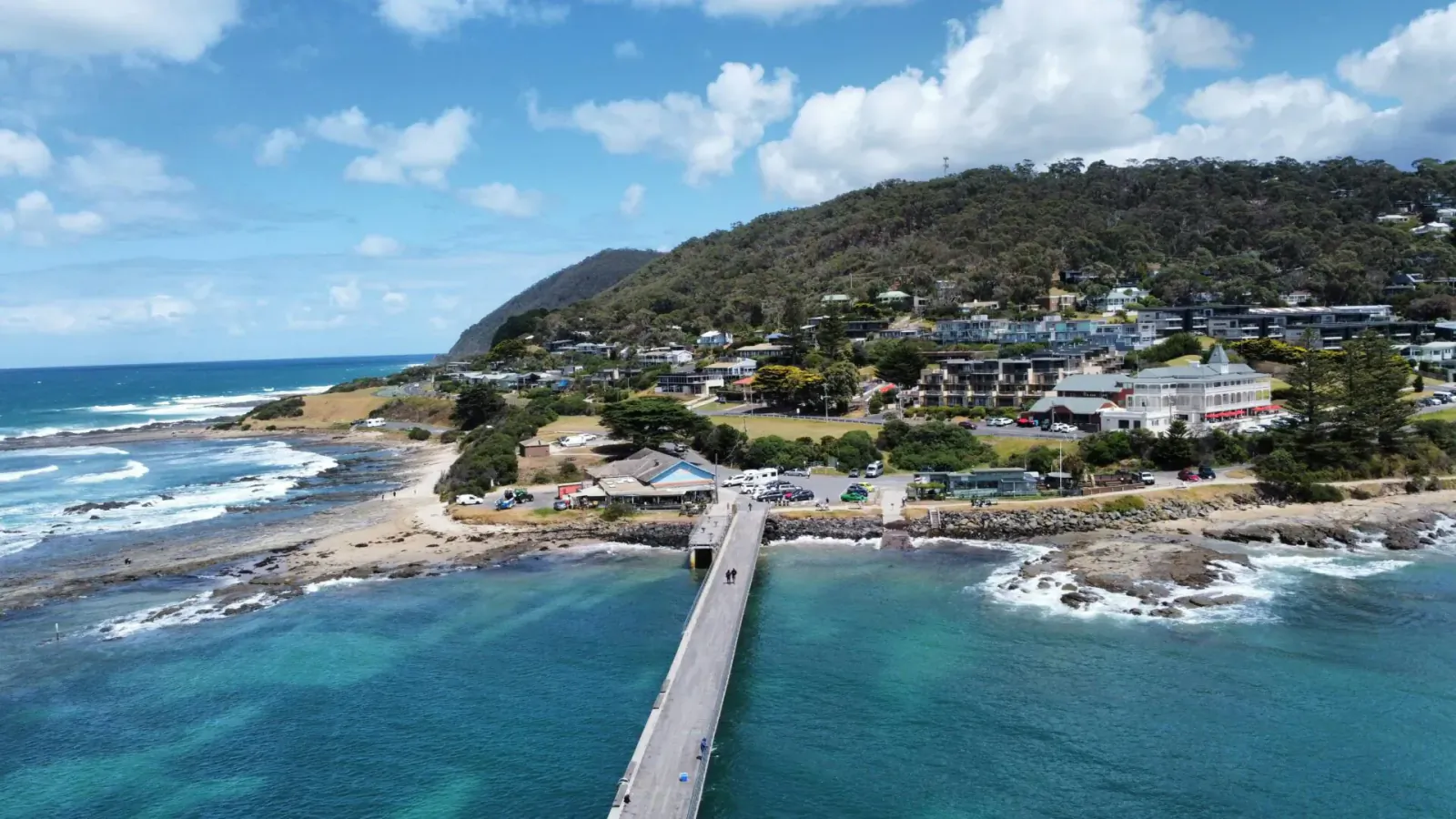 Aerial view of a long pier extending over turquoise ocean to a rocky beach with green hills and houses under blue sky.