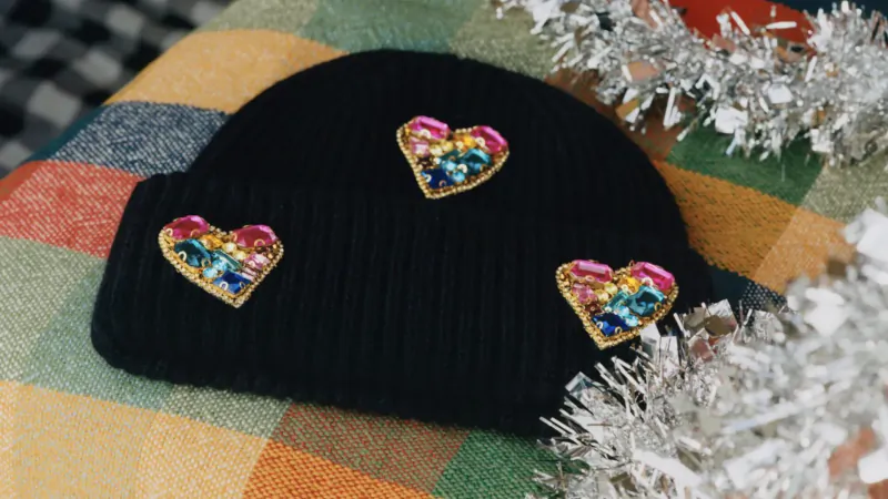 Black beanie hat with three sparkling rainbow heart patches on colorful plaid fabric with tinsel.