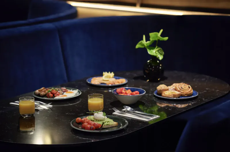 Luxurious hotel table with avocado toast, eggs royale, strawberries, pastries, juice on black glossy surface by blue velvet booth and plant.
