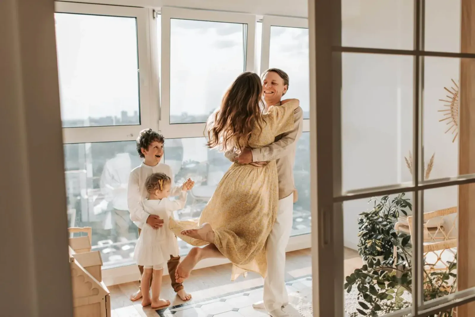 Family hugging joyfully by large windows in sunlit home, mom in yellow dress lifted by dad, kids smiling