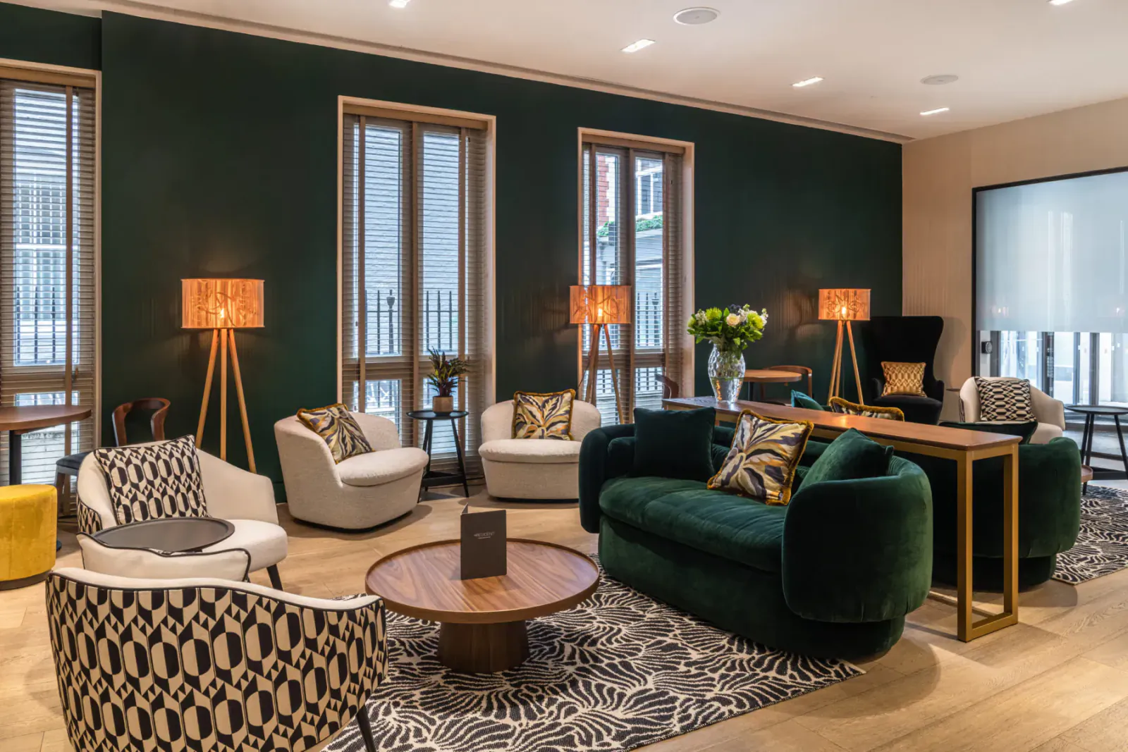 Modern hotel lounge with green velvet sofa, patterned chairs, wooden coffee table, lamps, and plants on hardwood floor.