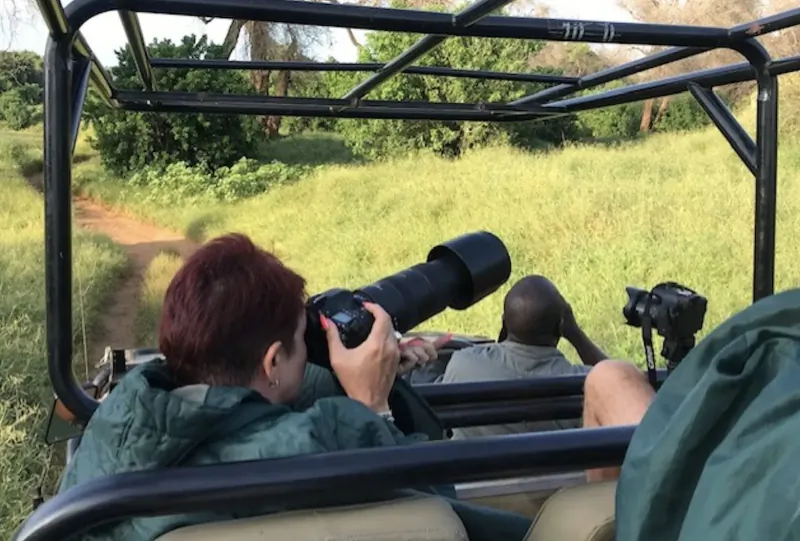 Woman with red hair and Black man photographing wildlife from open safari jeep in grassy savanna.