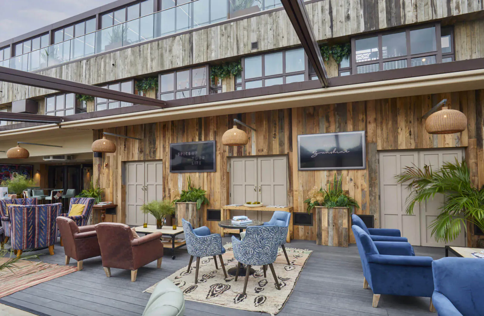 Cozy outdoor lounge with wooden walls, rattan lamps, blue armchairs, plants, and tables on deck under covered patio