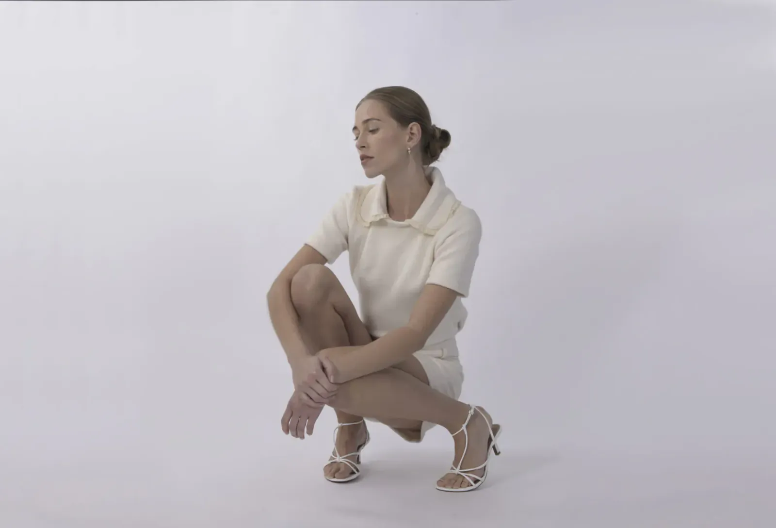 Woman in white short-sleeved dress and strappy heels squats thoughtfully on white background