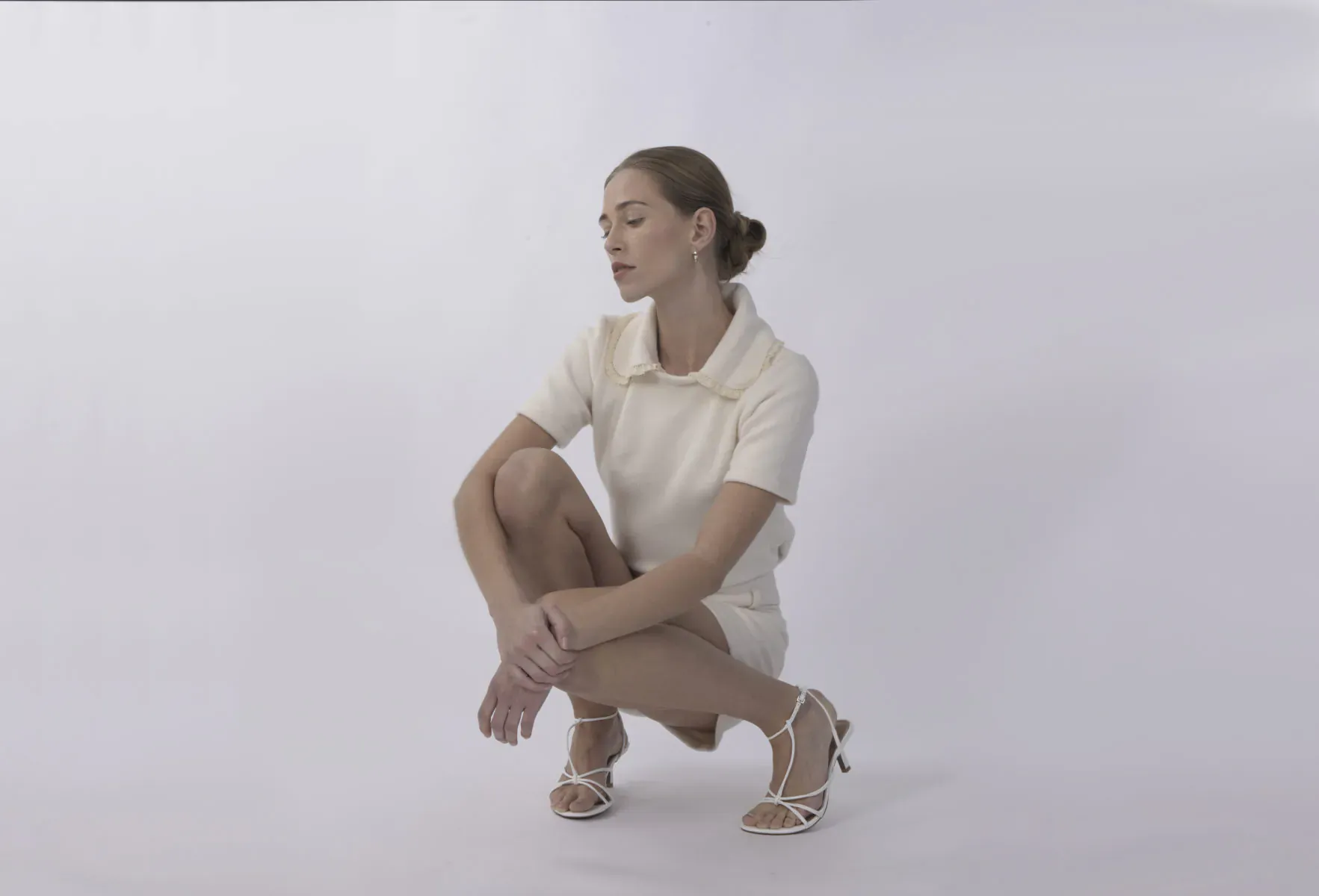 Woman in white short-sleeved dress and strappy heels squats thoughtfully on white background