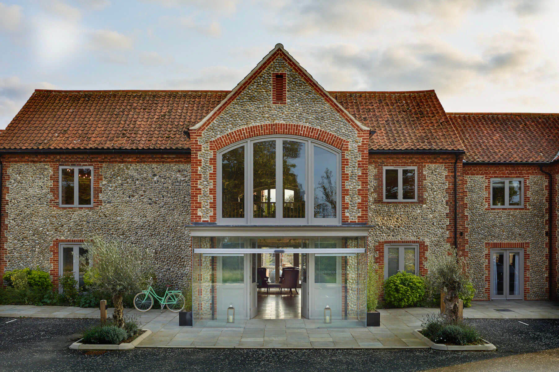 The Harper Norfolk: Stone barn-style country house with arched windows, glass entrance, olive trees, and bike on gravel forecourt at dusk.