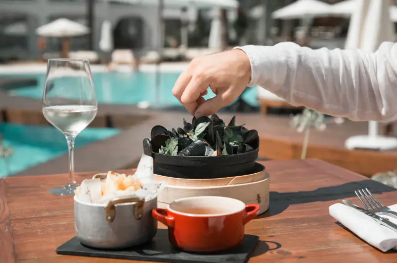 Hand pinching mussels in black steamer on wooden table with white wine, small bowls, poolside at Destino Pacha Ibiza