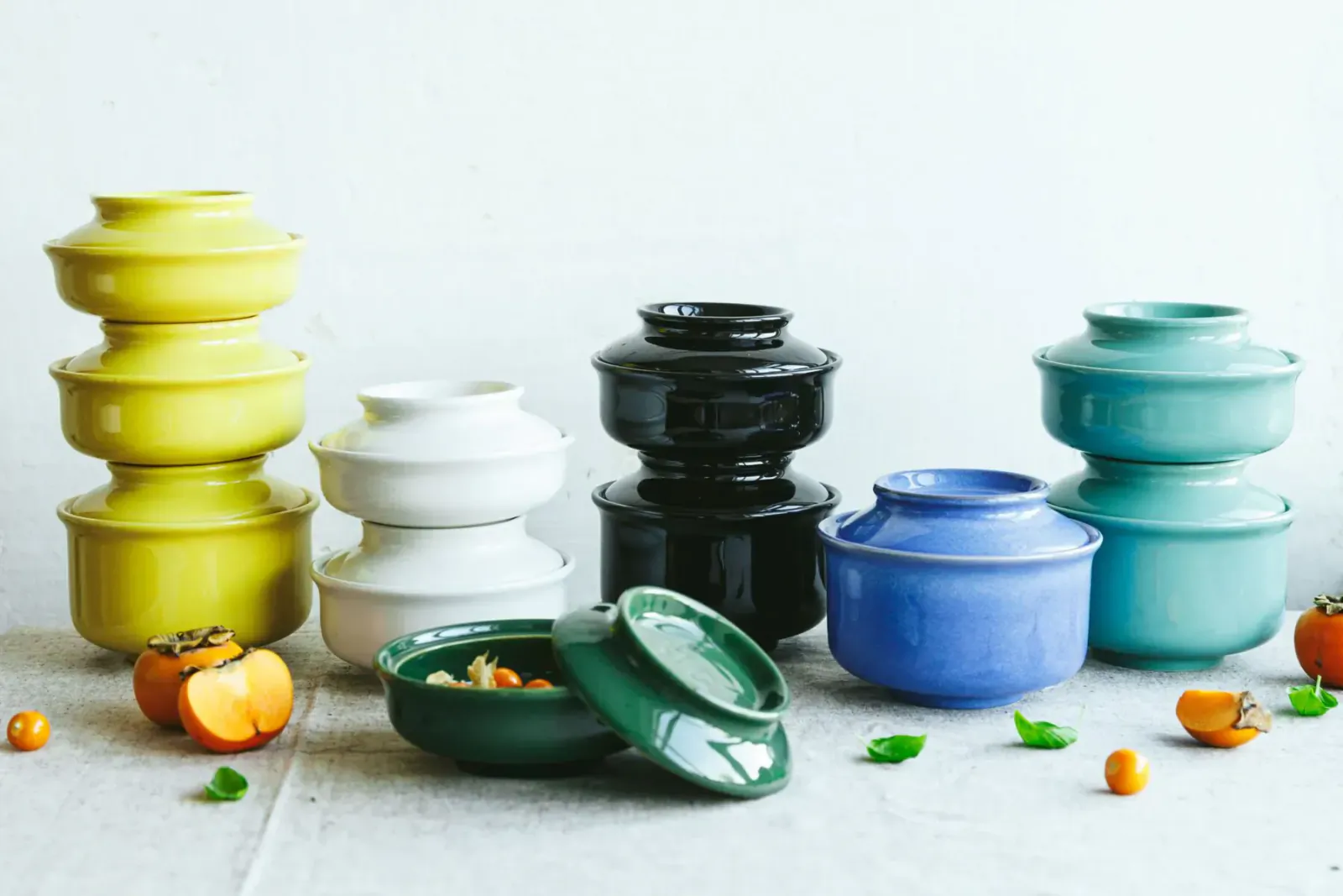 Assortment of Hokan stacking bowls in yellow, white, black, green, blue, teal with lids, some with food, persimmons nearby.