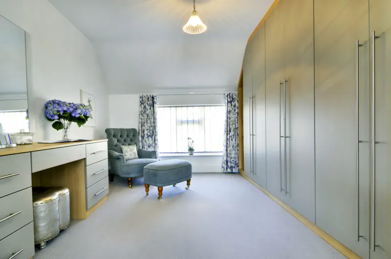 Luxurious cottage dressing room with curved grey wardrobes, white vanity, blue hydrangeas, tufted chair, and ottoman.